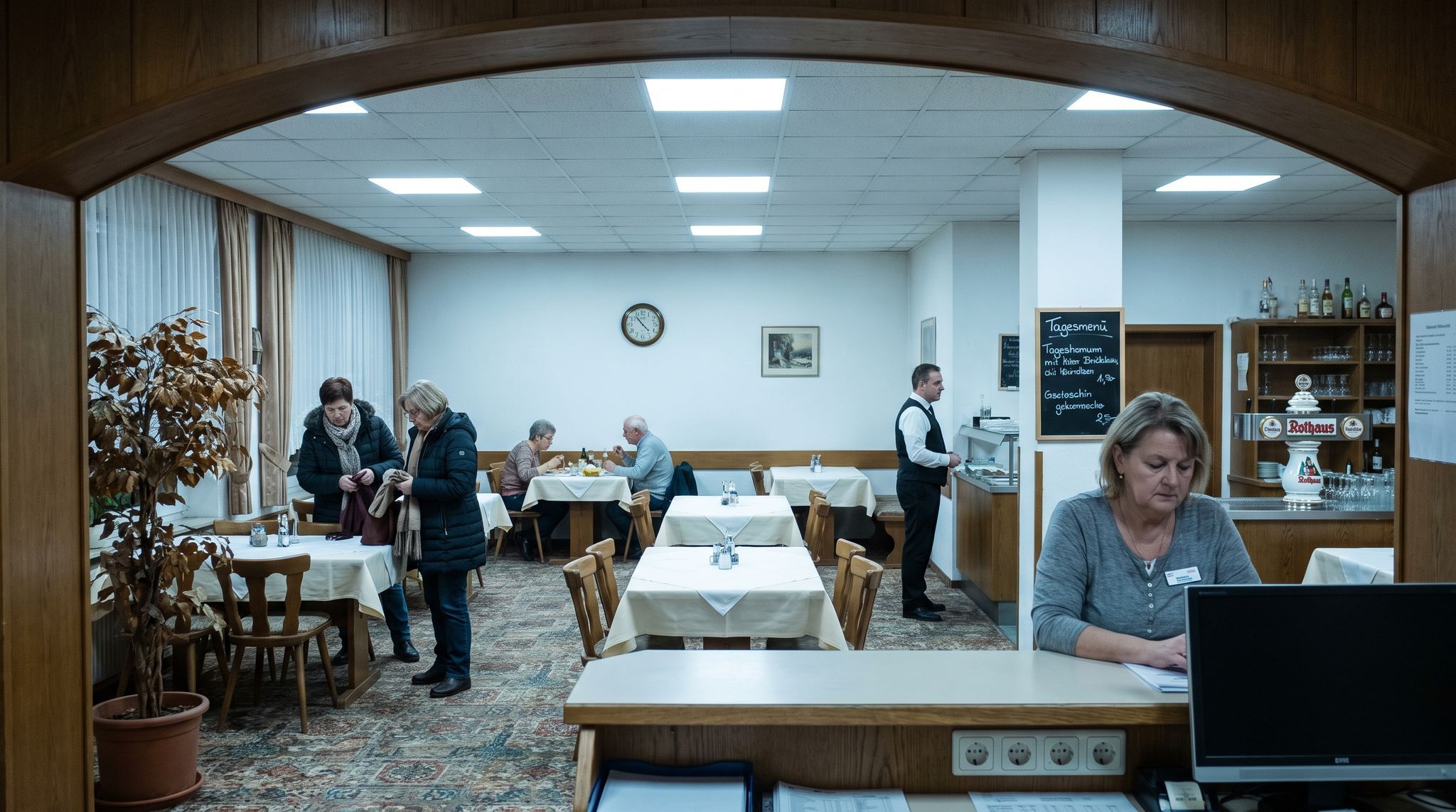 The view from a hotel lobby into a two-thirds empty restaurant at evening service, under flat overhead light: one couple eating in silence, two more putting their coats on to leave, and a waiter standing still with his order pad